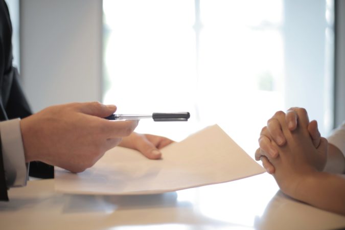 cropped image of one person handing a pen and small stack of papers across a table to another person with folded hands