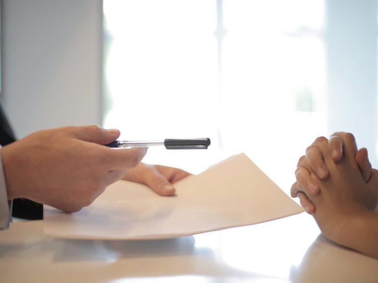 cropped image of one person handing a pen and small stack of papers across a table to another person with folded hands