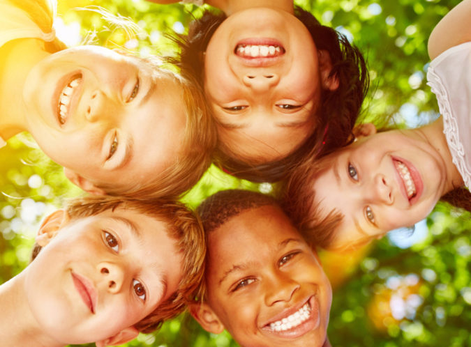 children standing in a circle with their heads touching smiling down at the camera under tree leaves on a sunny day
