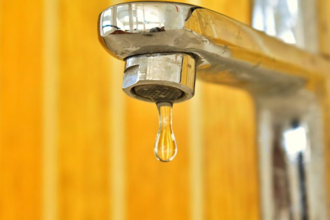 Close up of water dripping out of a faucet with a yellow background