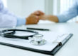 clip board and stethoscope in focus in the foreground, doctor holding a patient's hand out of focus in the background
