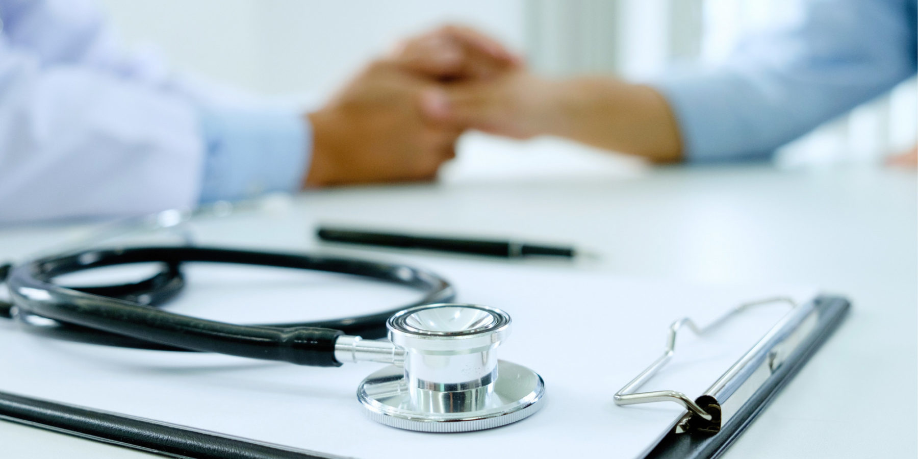 clip board and stethoscope in focus in the foreground, doctor holding a patient's hand out of focus in the background