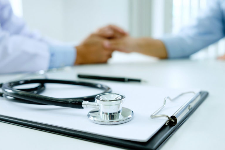 clip board and stethoscope in focus in the foreground, doctor holding a patient's hand out of focus in the background