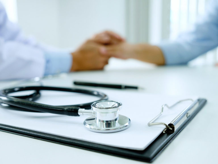 clip board and stethoscope in focus in the foreground, doctor holding a patient's hand out of focus in the background
